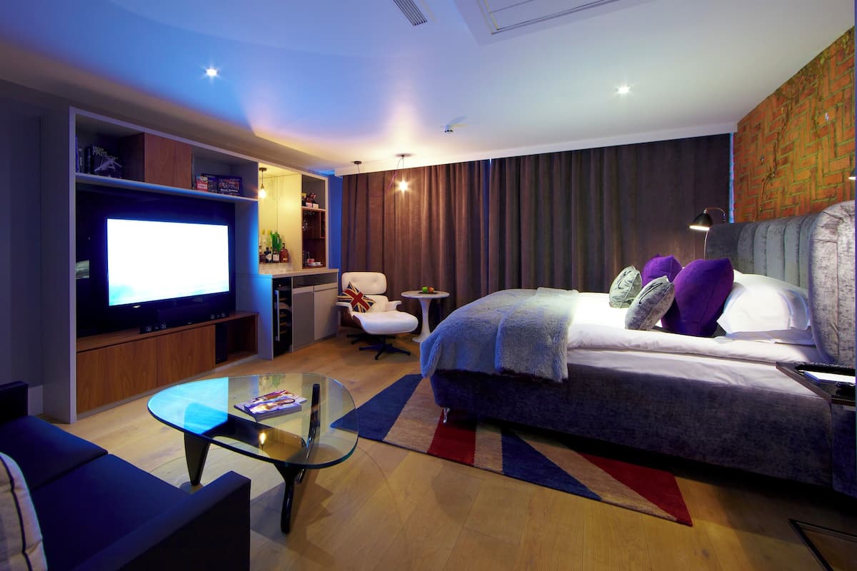 A modern hotel room with a large flat-screen TV, a comfortable bed with multiple pillows, a small round table, and a seating area with a sofa and armchair.