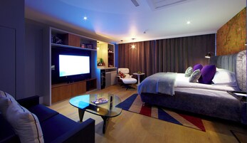 Ein modernes Hotelzimmer mit großem Flachbildfernseher, einem bequemen Bett mit vielen Kissen, einem kleinen runden Tisch und einer Sitzecke mit Sofa und Sessel.