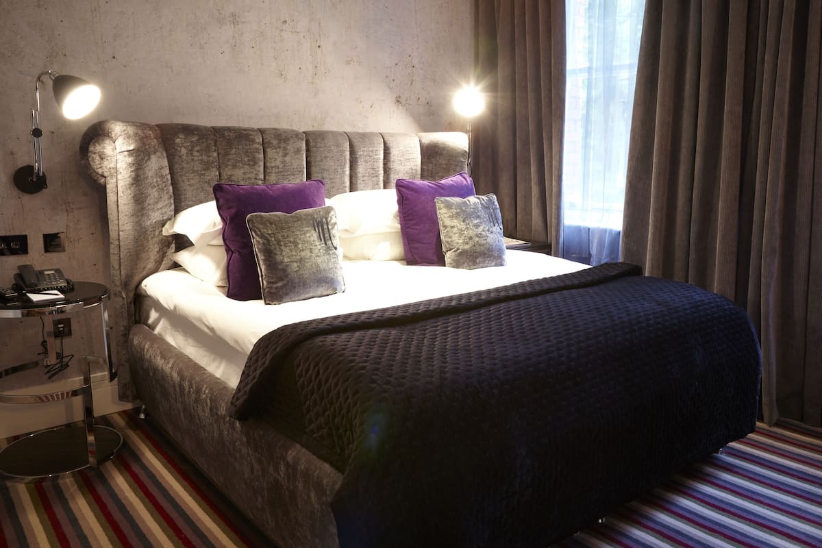A bed with a grey headboard, white bedding, and purple pillows. There are two bedside lamps, a striped carpet, and a window with curtains.