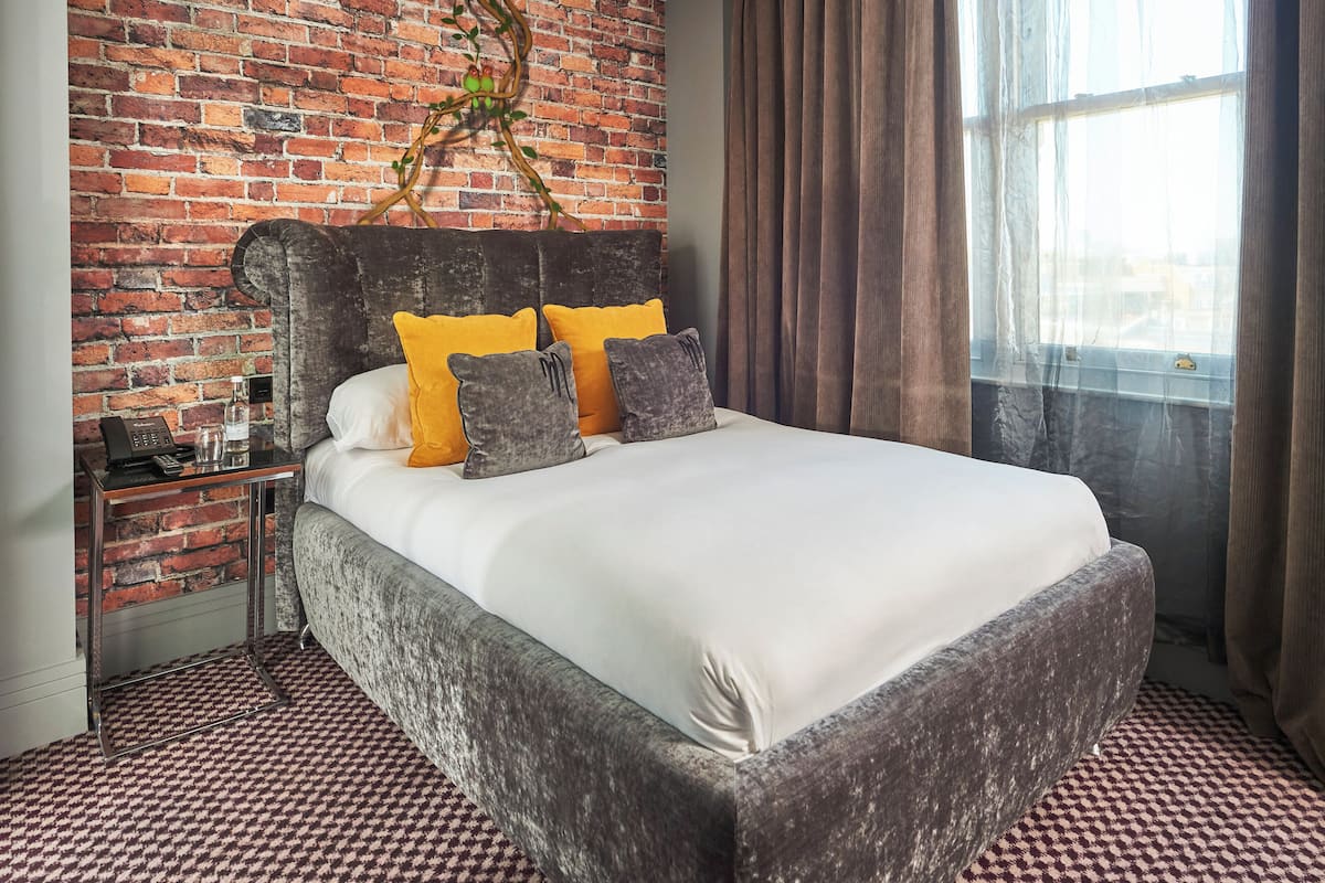 A bed with a dark headboard, white bedding, and two pillows, one yellow and one gray, in a room with a brick wall and a window with curtains.