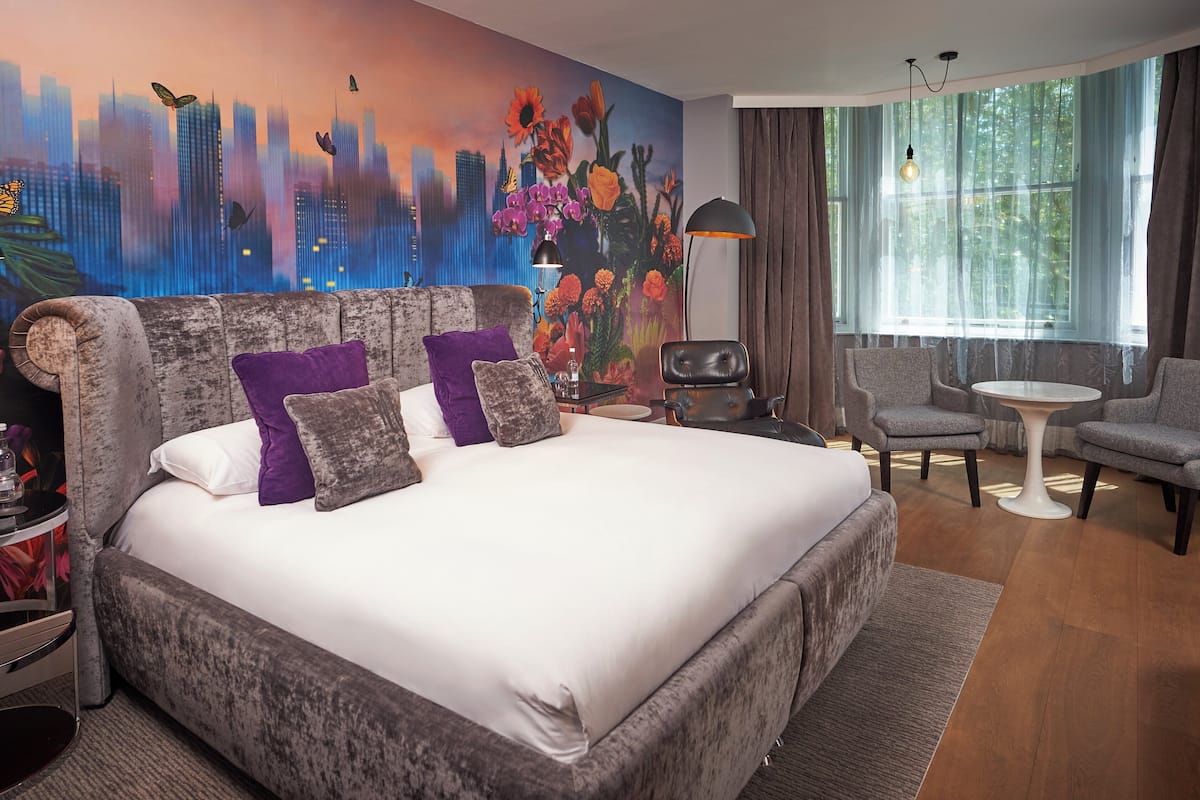 A hotel room with a large bed, two armchairs, a small table, and a cityscape mural on the wall.