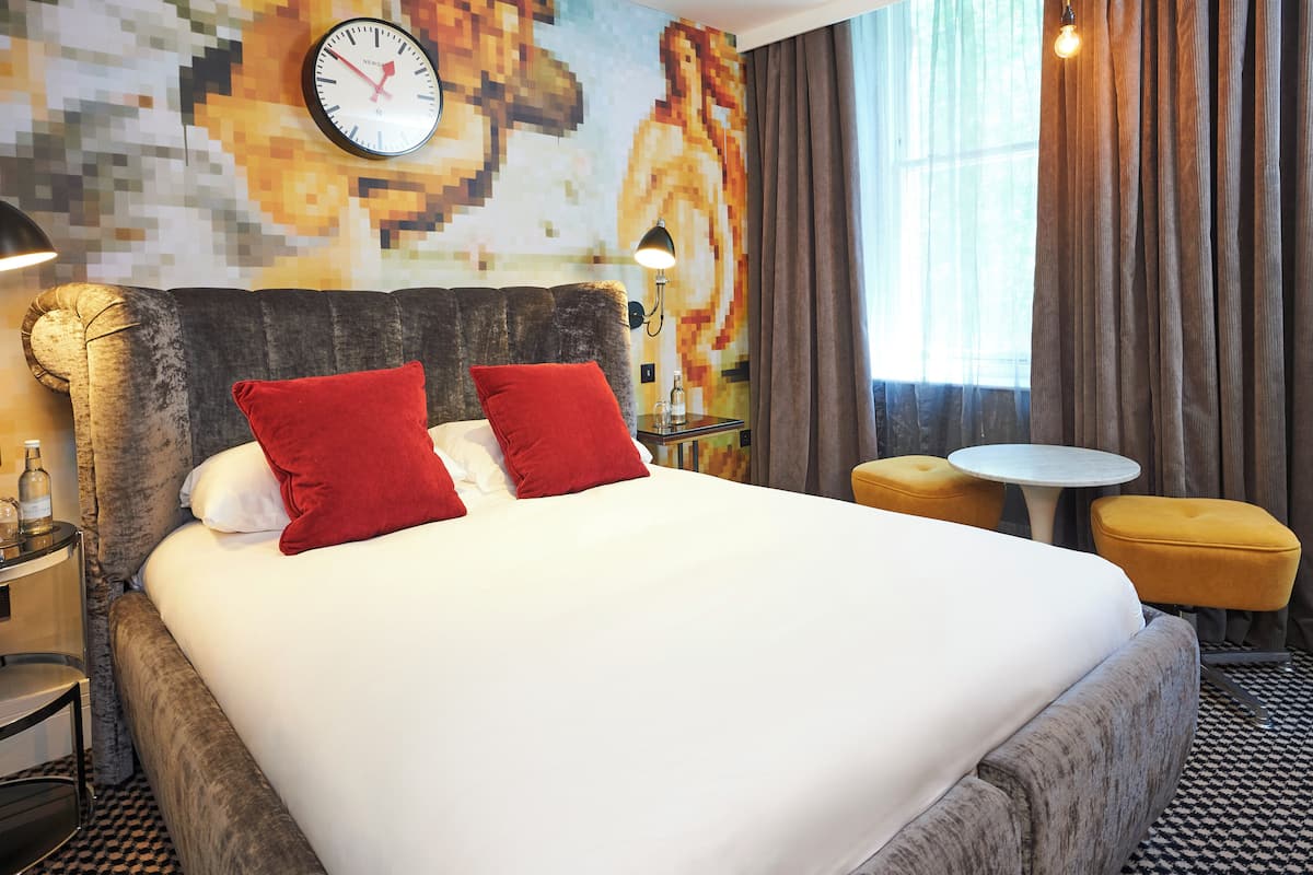 A neatly made bed with red pillows, a wall clock, a lamp, and a small round table with a yellow ottoman.