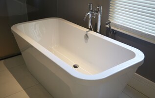 Ein modernes Badezimmer mit einer freistehenden Badewanne, einer Handbrause und einem Fenster mit Jalousien.