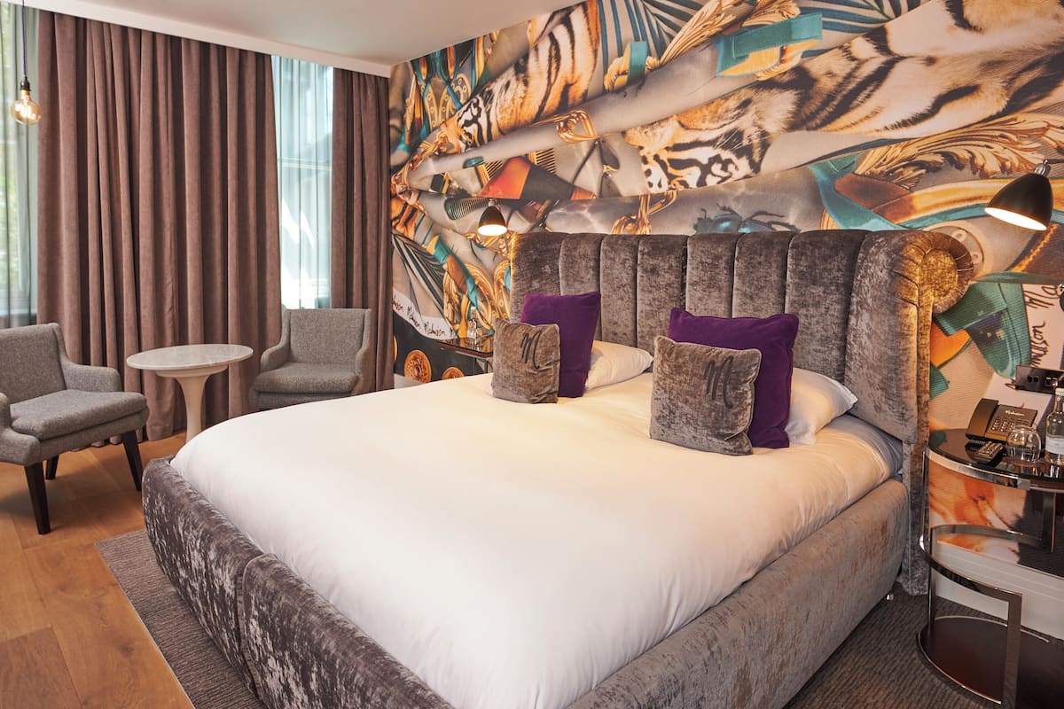 A hotel room with a large bed, two armchairs, a small round table, and a decorative headboard.