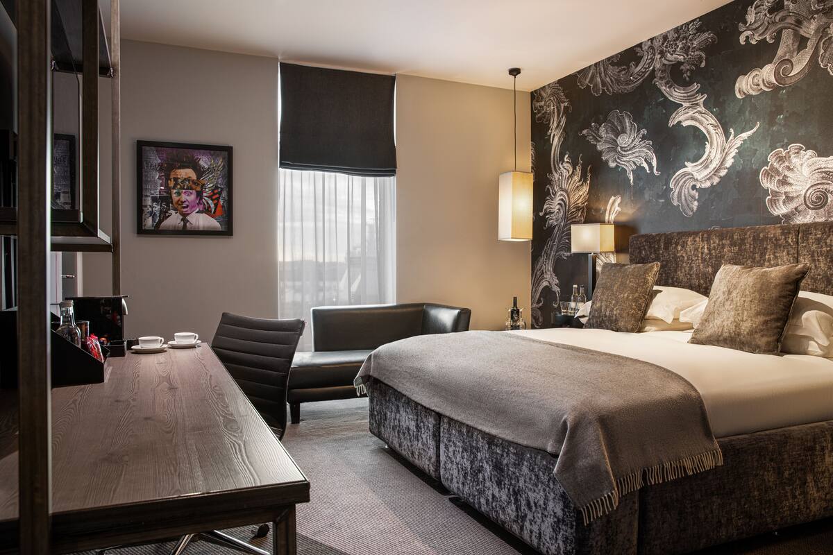 A hotel room with a large bed, a desk with a chair, a window with sheer curtains, and a decorative wall with a floral pattern.
