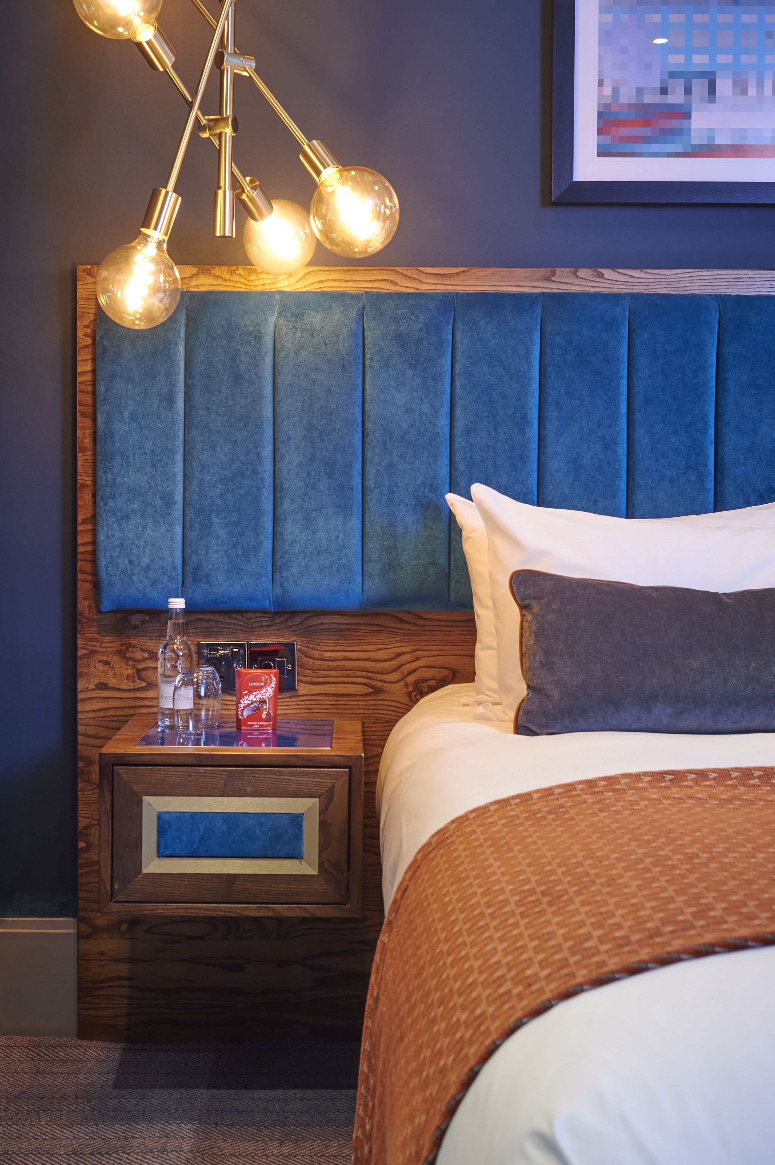 A hotel room with a wooden nightstand, a blue upholstered headboard, a bed with white and brown bedding, and a wall-mounted light fixture.