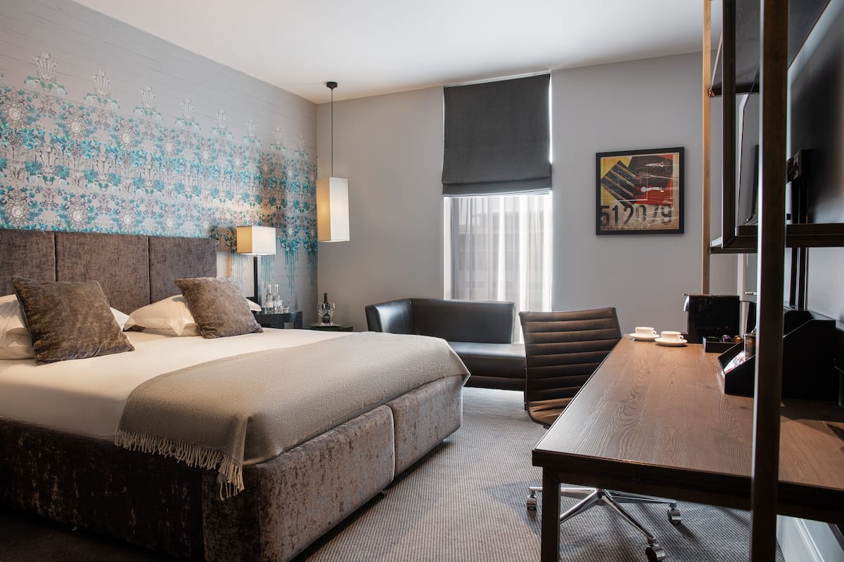 A hotel room with a large bed, a desk, a chair, a window with curtains, a wall with floral wallpaper, and a framed picture on the wall.