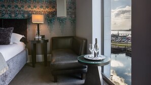 A hotel room with a bed, a leather armchair, a glass table with two wine glasses, a lamp, and a view of a body of water through a window. - Malmaison Liverpool (Liverpool)