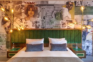 Una habitación de hotel con un cabecero tapizado en verde, una cama grande con ropa de cama blanca y gris, dos mesitas de noche con lámparas, y una pared decorada con carteles y una impresión de gran formato.