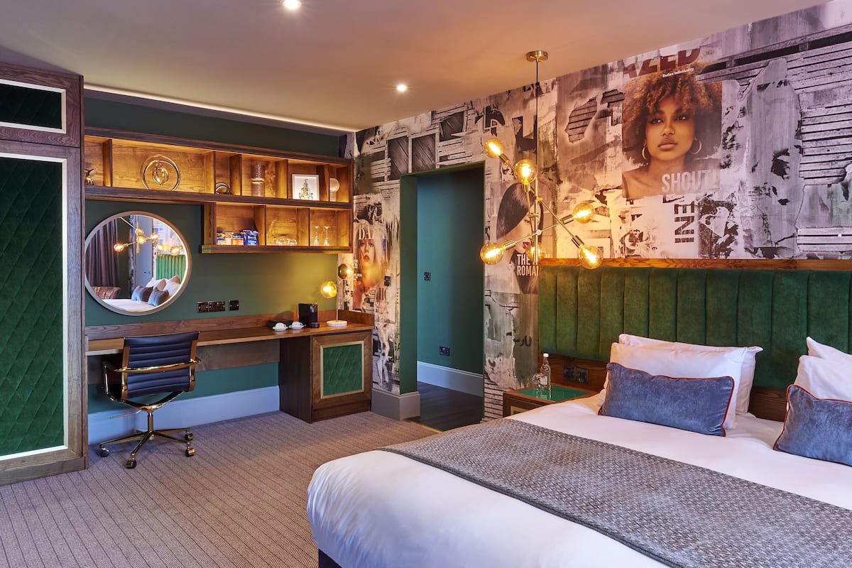A hotel room with a bed, a desk, a chair, a mirror, and a wall with a poster.