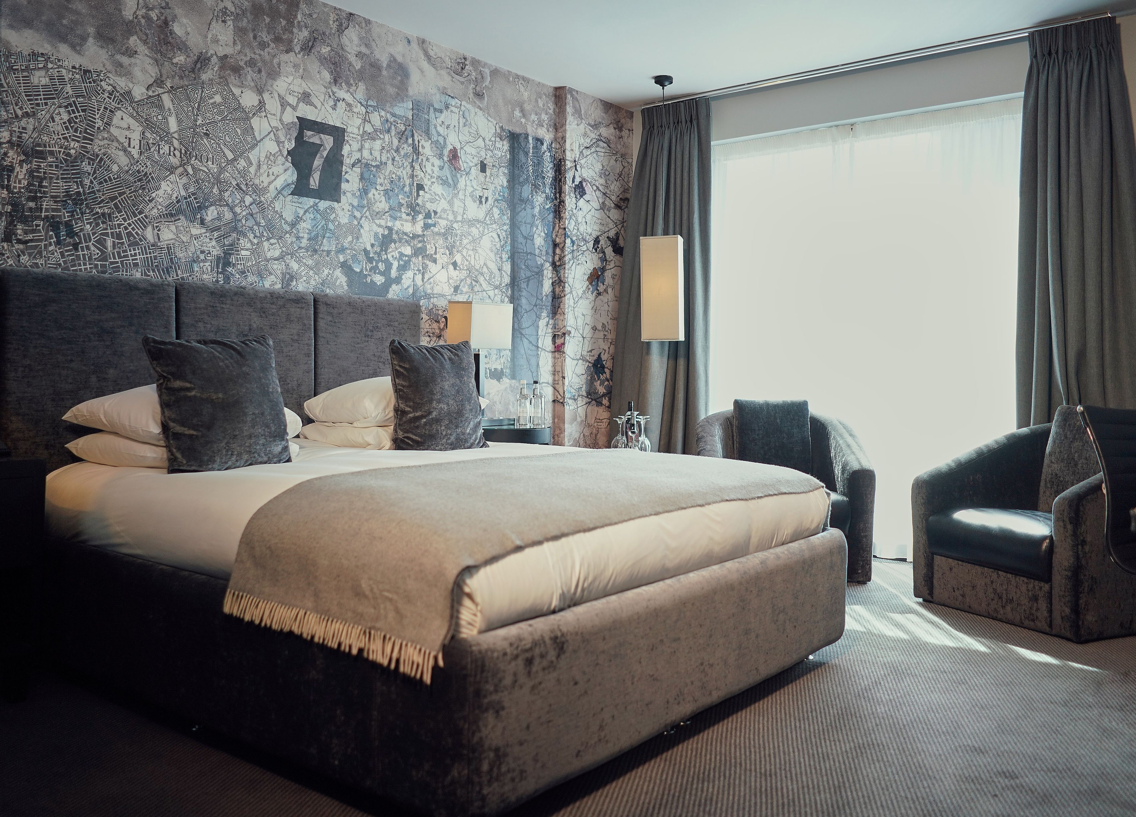 Habitación de hotel con una cama grande, cabecera, mesita de noche, lámpara, una silla y un ventanal con cortinas.