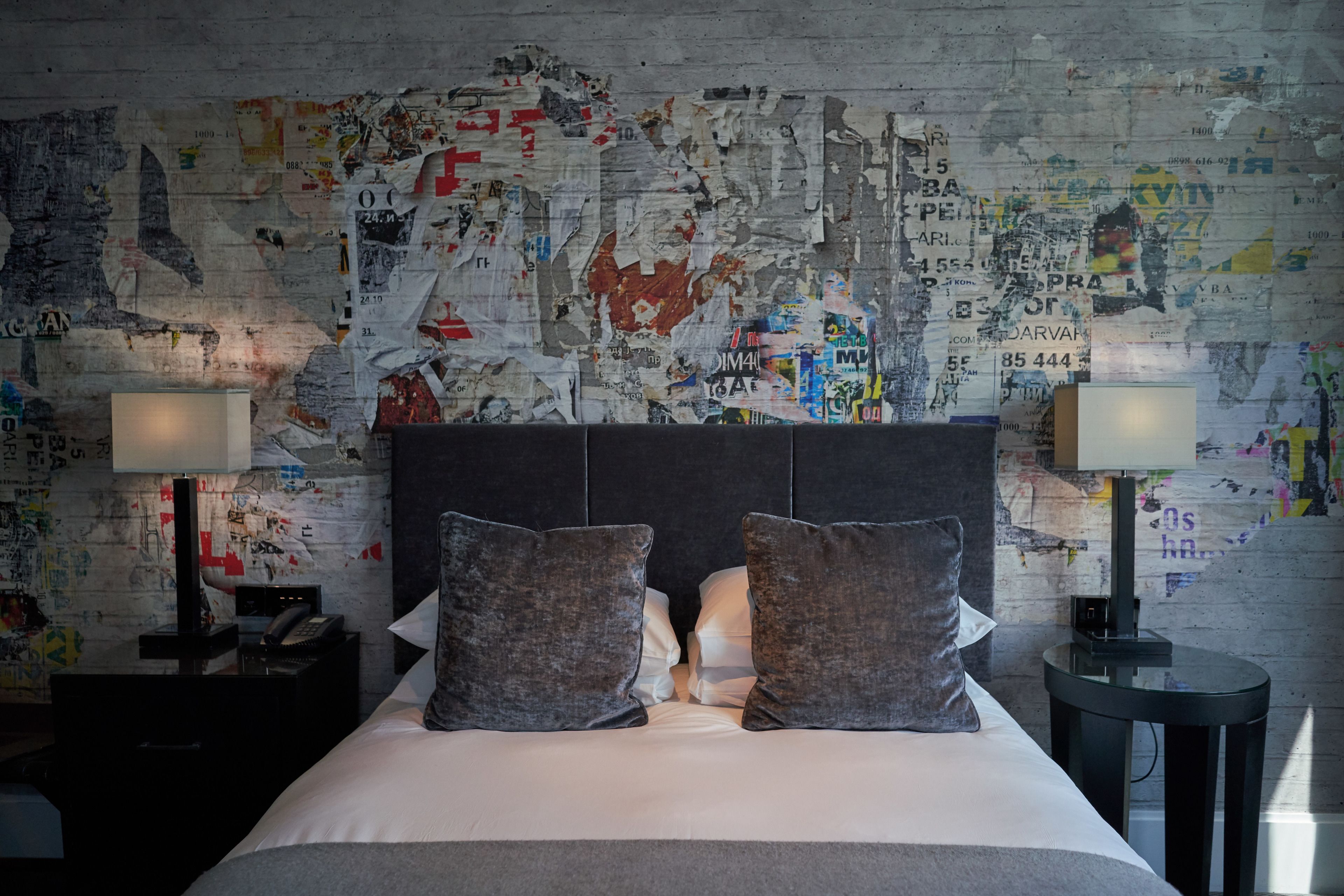 Une chambre d’hôtel avec un lit, deux oreillers, une table de chevet avec une lampe, et un mur recouvert de divers affiches et autocollants.