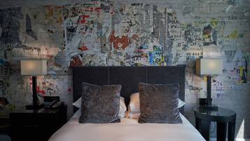 Ein Hotelzimmer mit einem Bett, zwei Kissen, einem Nachttisch mit Lampe und einer Wand voller verschiedener Poster und Aufkleber.