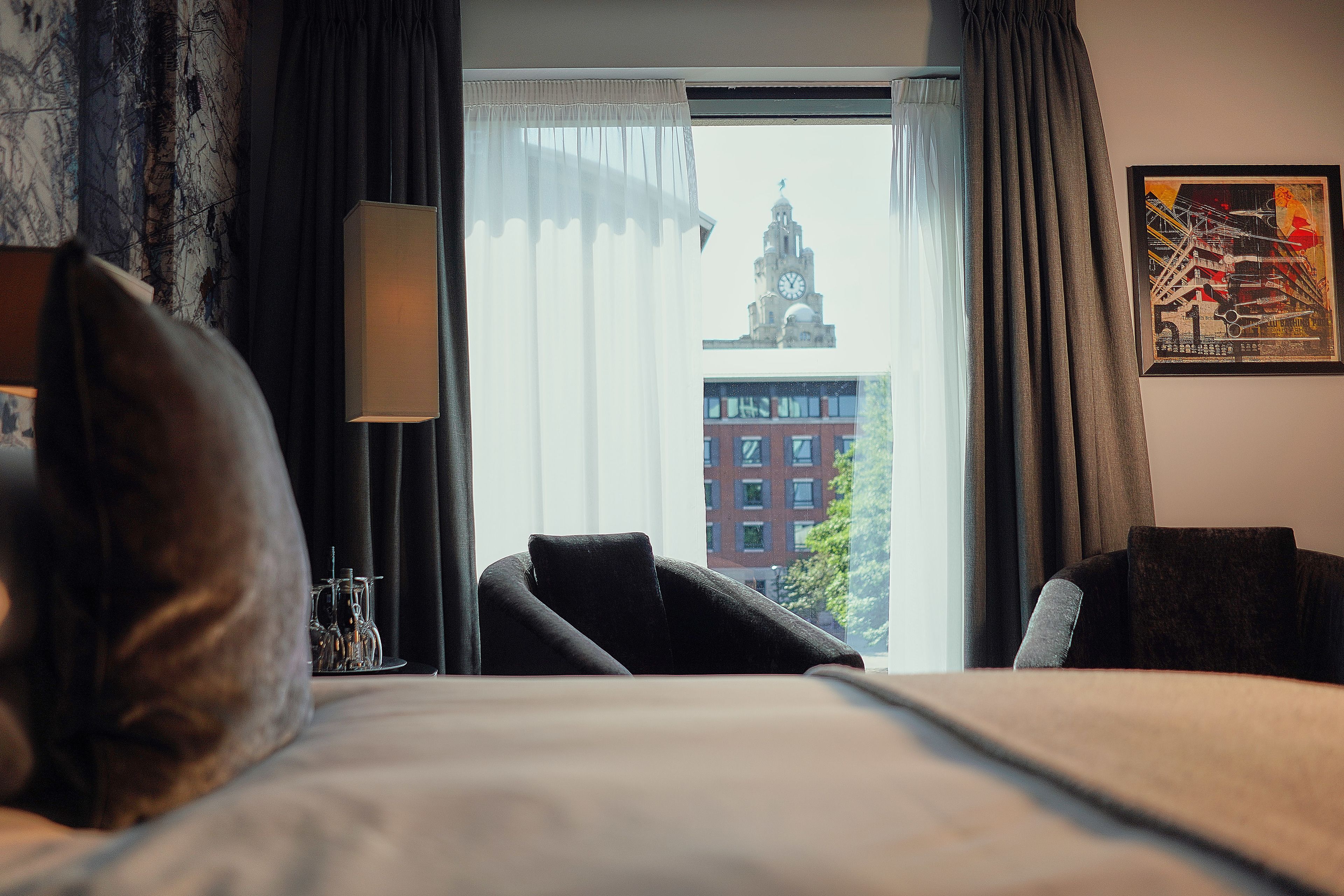Quarto de hotel com cama, uma cadeira, abajur e vista para um edifício com torre de relógio pela janela.