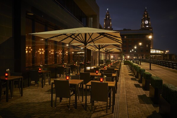 An outdoor dining area with tables and chairs, a large umbrella, and a building with a clock tower in the background. - Malmaison Liverpool (Liverpool)