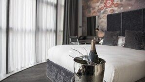Habitación de hotel con cama, un ventanal con cortinas, un adorno de pared y un minibár con una botella y vasos.