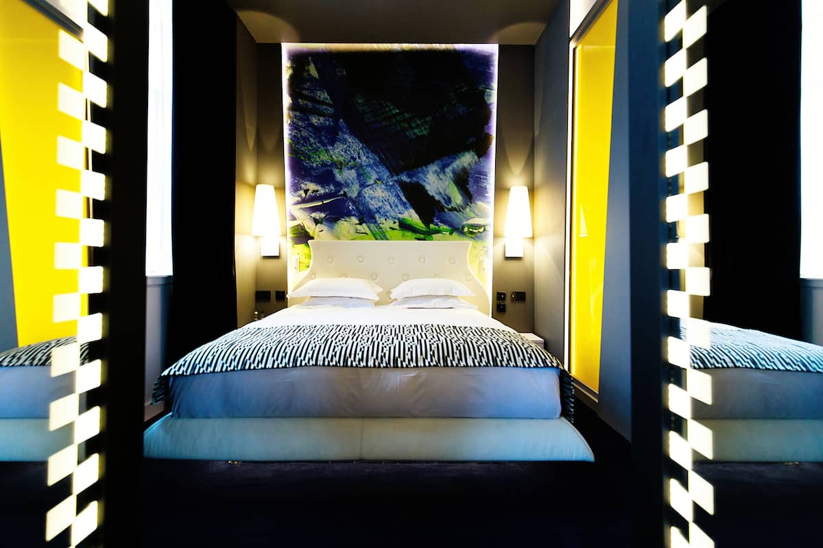 A modern hotel room with a large bed, two bedside lamps, and a large abstract painting on the wall.