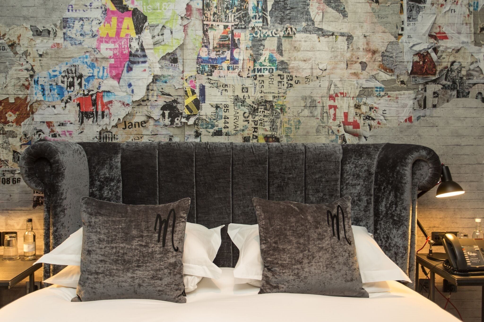 A bed with a headboard covered in various posters and stickers, a bedside table with a lamp, and a wall with a map-like design.