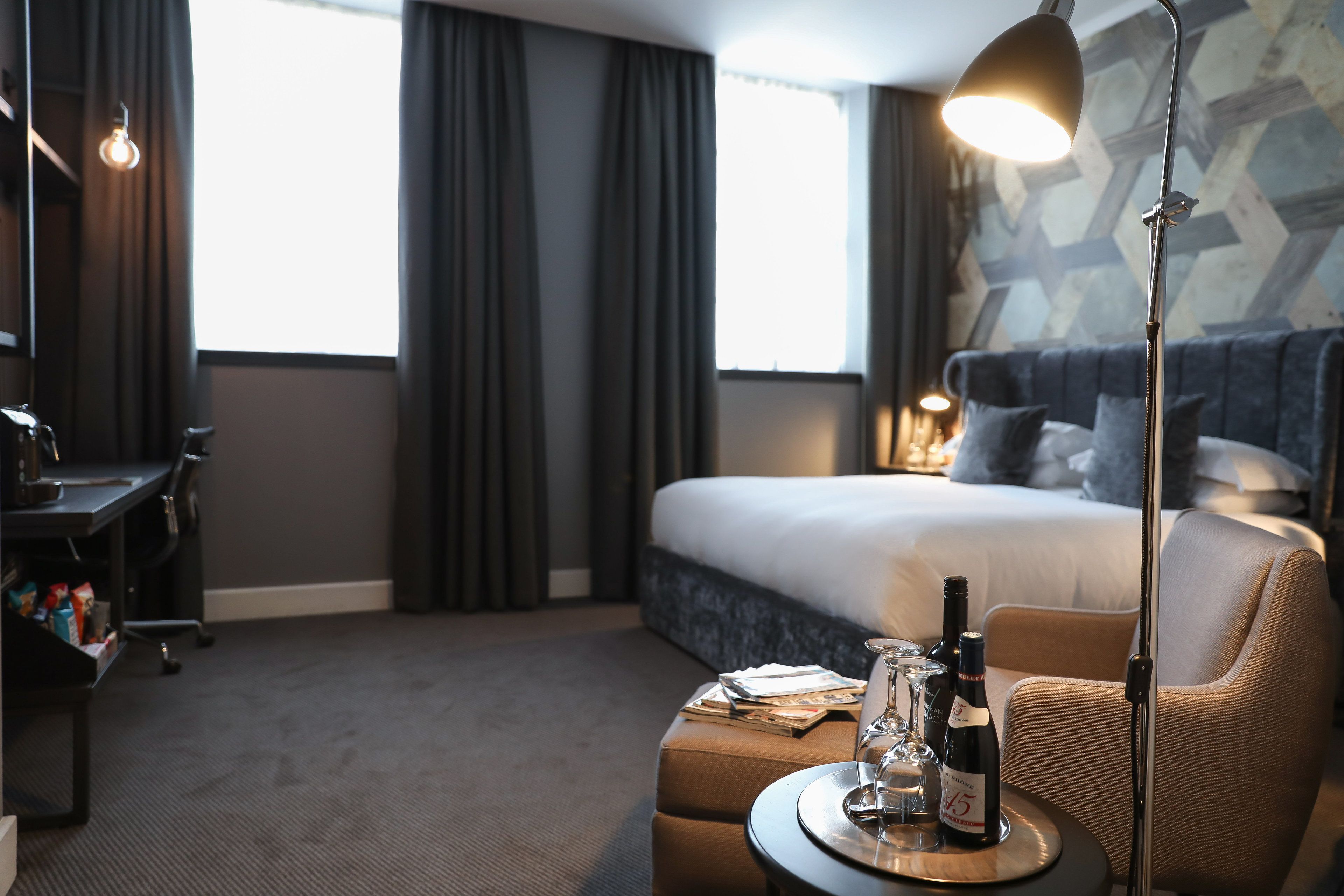 A hotel room with a bed, a desk, a chair, a table with a bottle and glasses, and a floor lamp.