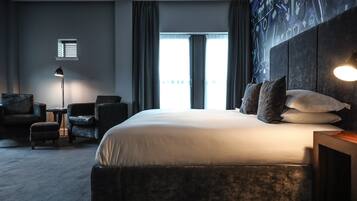 A modern hotel room with a large bed, dark headboard, and a view of the city through large windows.