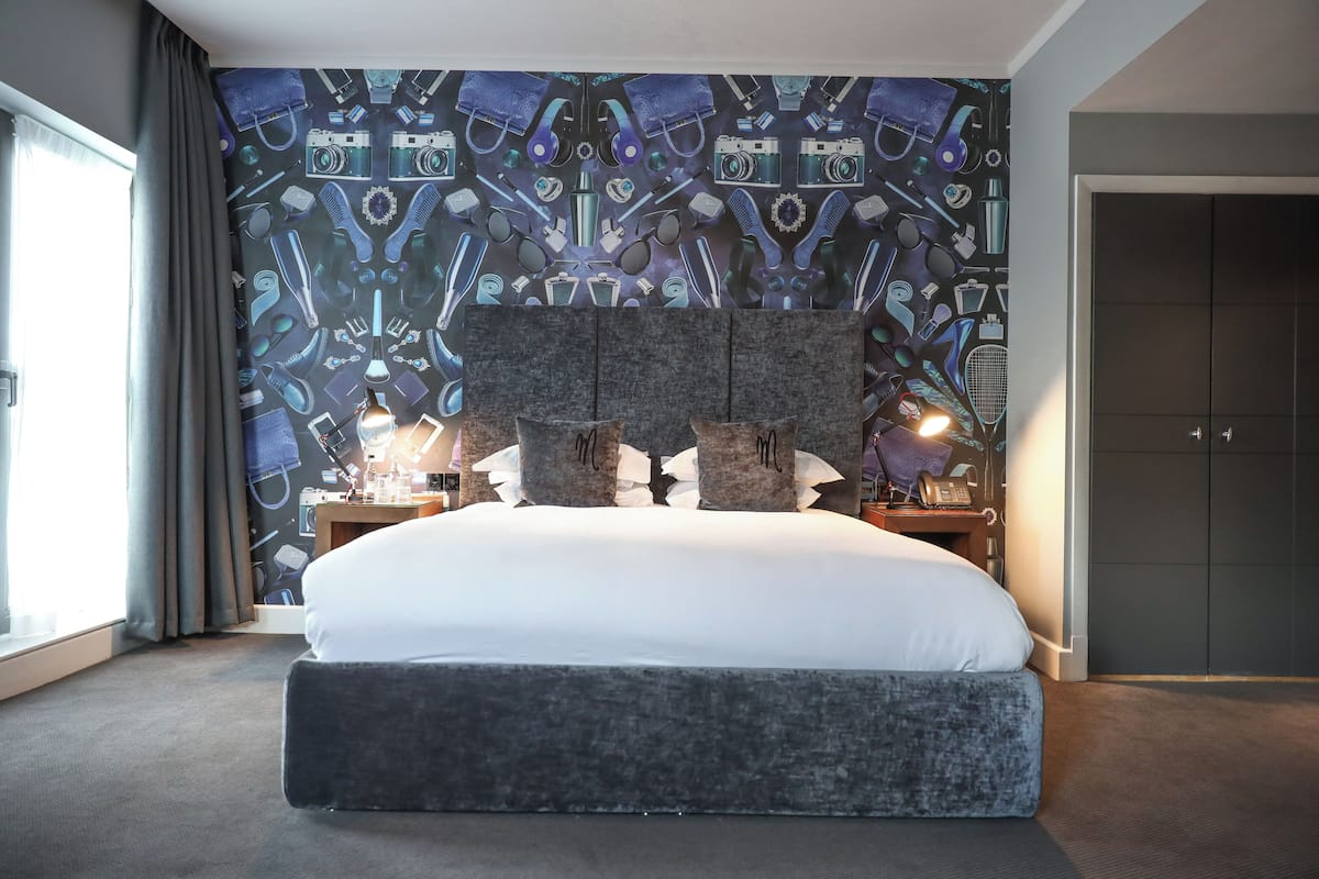 A bedroom with a large bed, a headboard, bedside lamps, a nightstand, and a wall with a pattern of various items.