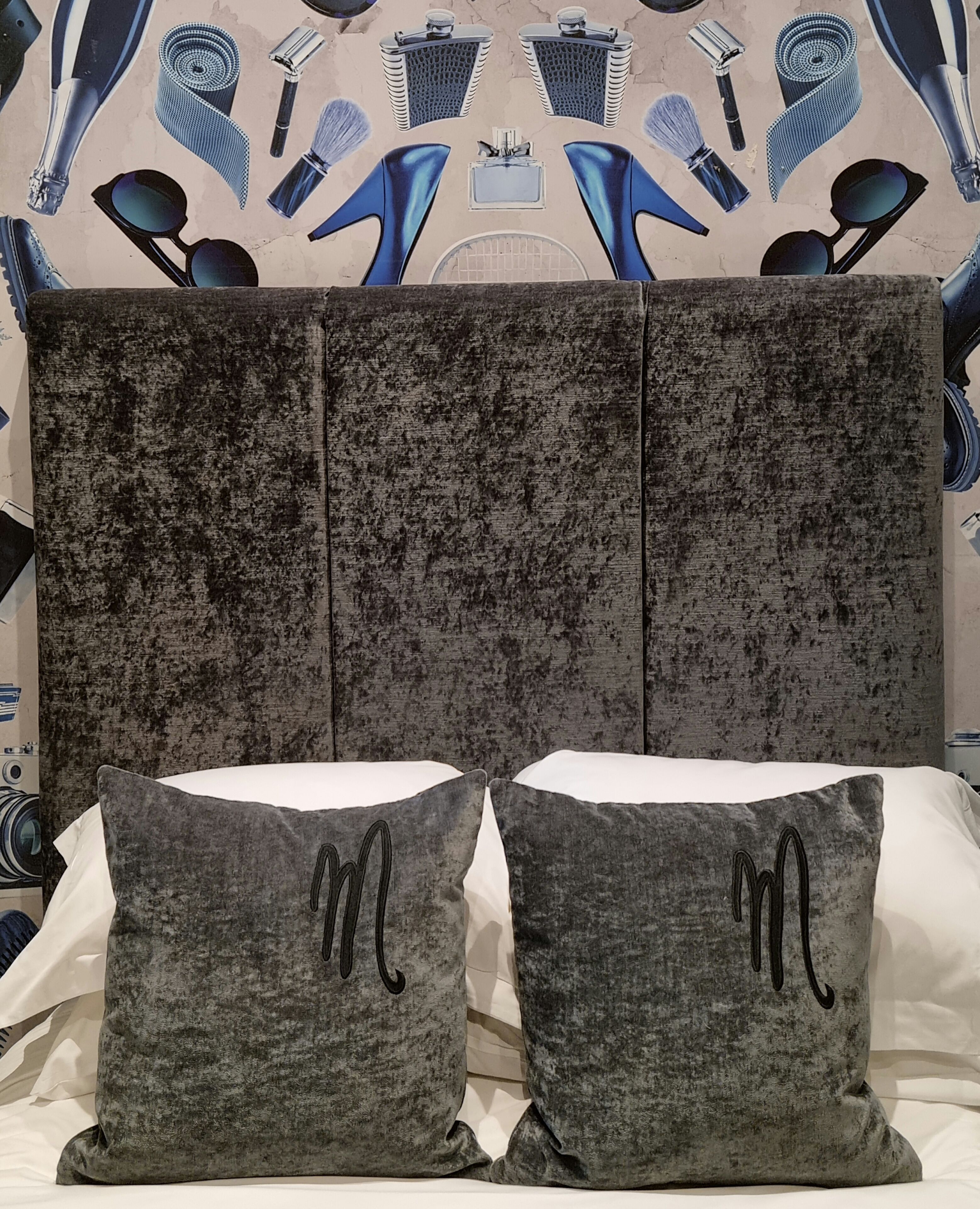 A bed with grey pillows featuring a monogram, a textured grey headboard, and a wall with a patterned design.