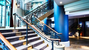 Un moderno vestíbulo de hotel con una escalera monumental, un ascensor de cristal y un área de descanso con una planta en maceta.