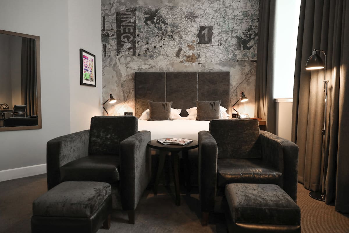 A hotel room with a large bed, two armchairs, a small table, and a desk with a lamp.
