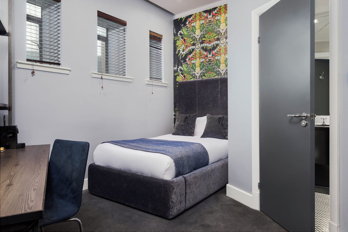 A bedroom with a bed, a desk, and a chair. There are windows with blinds, a door, and a wall with a colorful mural.