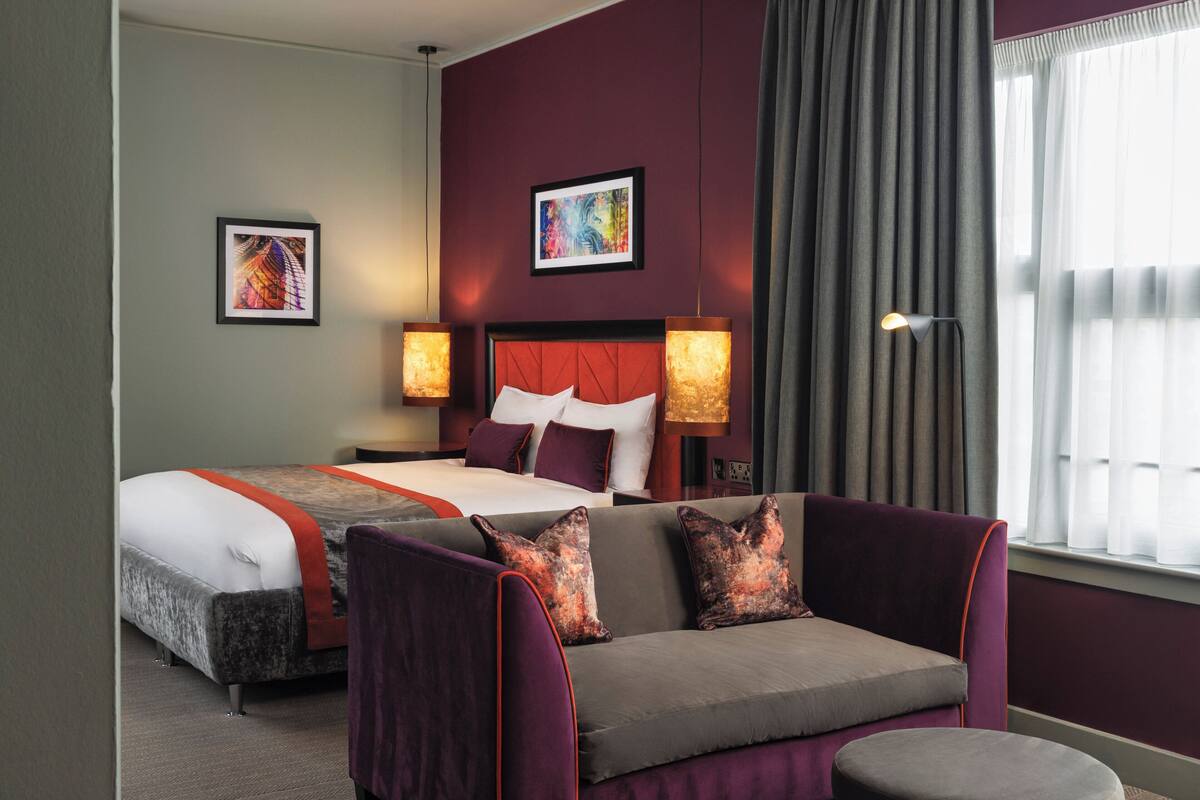 A hotel room with a bed, a sofa, two bedside lamps, and artwork on the walls.