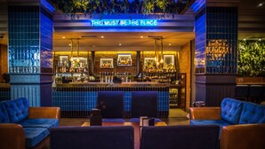 A bar with a sign reading "THIS MUST BE THE PLACE" and a variety of bottles on shelves. There are blue upholstered chairs and a bar counter. - Malmaison Newcastle (Newcastle-upon-Tyne)