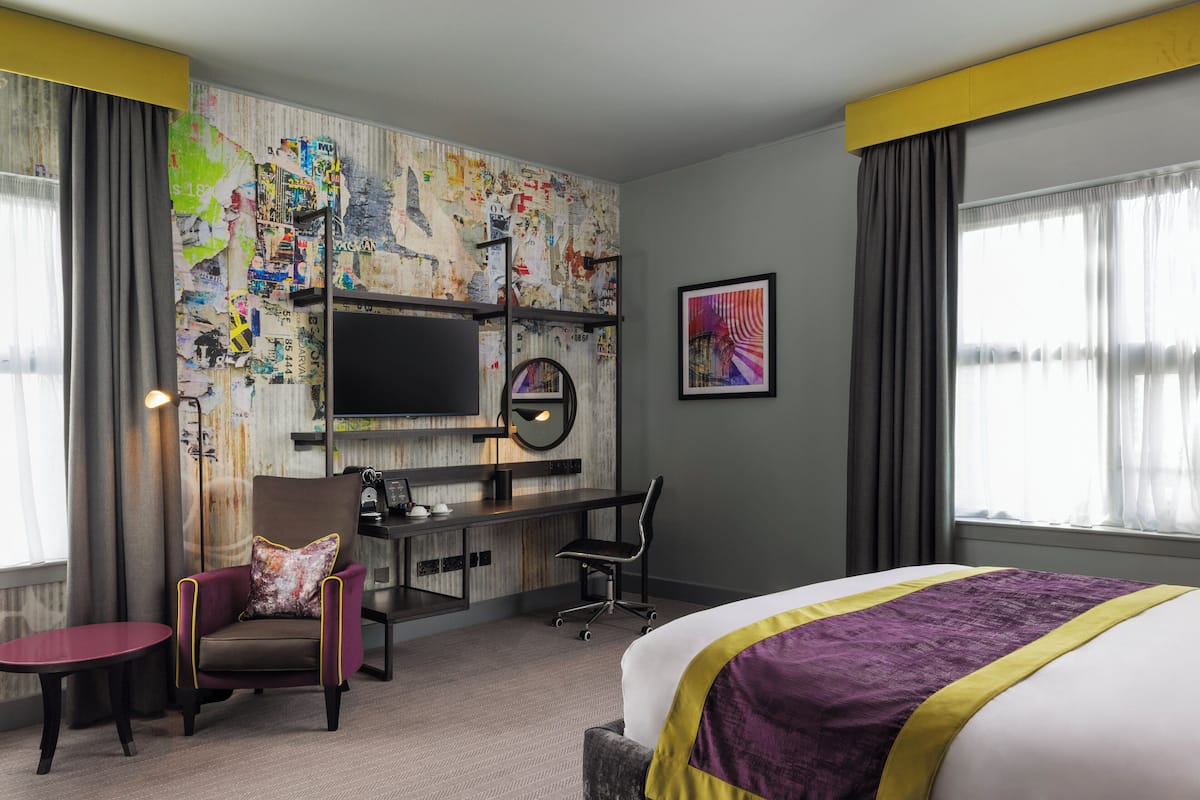 A hotel room with a bed, a desk with a computer, a chair, a TV, and a large window with curtains.