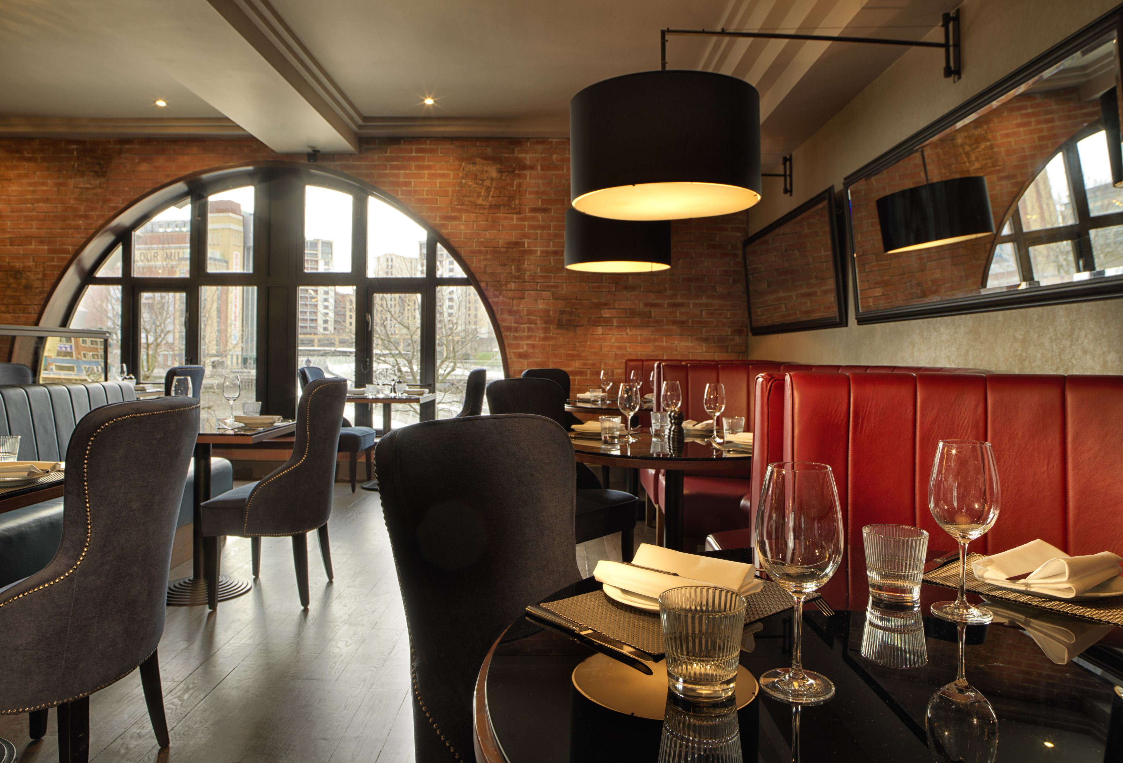 A restaurant with a brick wall, large windows, and a red booth. Tables are set with wine glasses and cutlery.