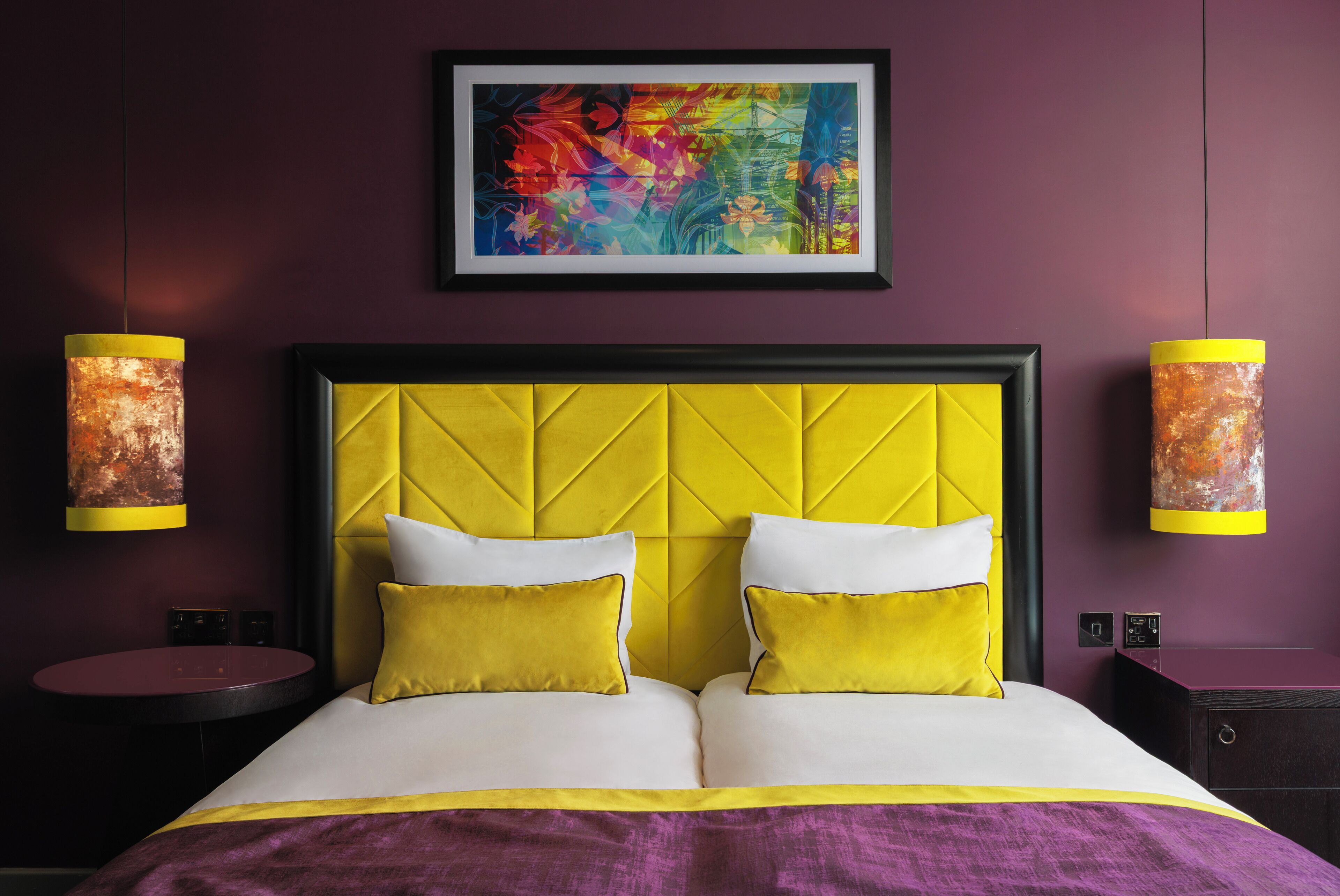 A hotel room with a yellow headboard, two yellow pillows, a purple bedspread, and a framed abstract painting above the bed.