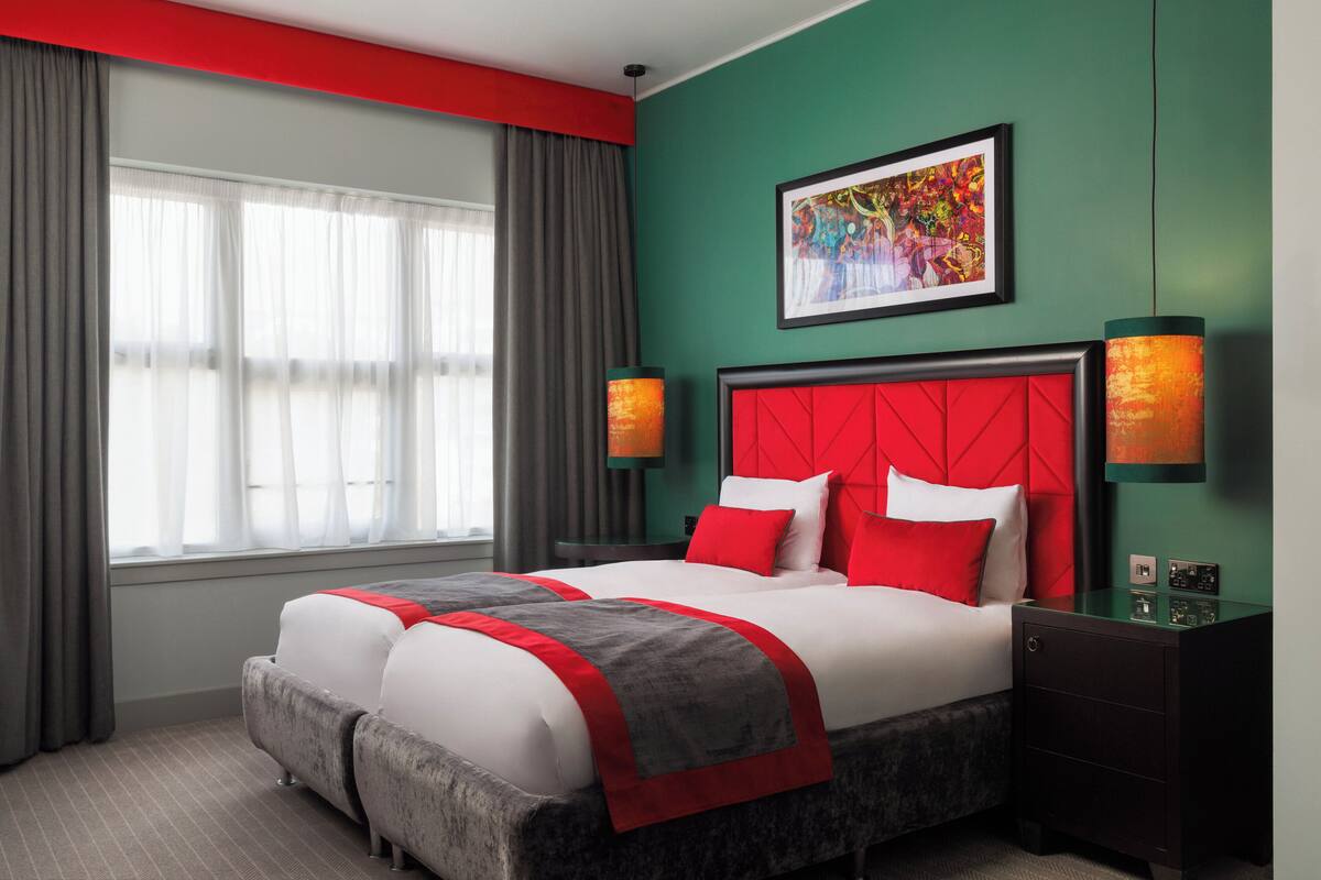 A hotel room with a large bed, grey and red bedding, a red headboard, two wall-mounted lamps, a green accent wall, and a window with grey curtains.