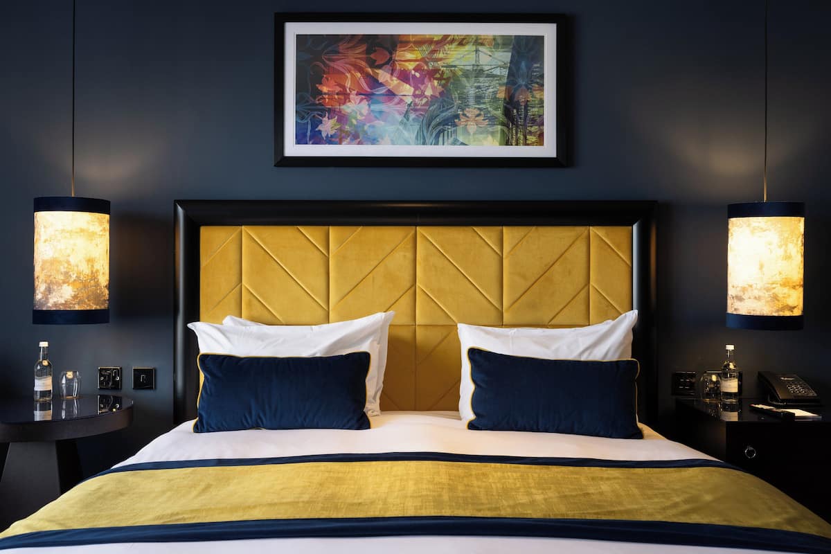 A hotel room with a large bed, a headboard with a geometric pattern, two bedside lamps, a framed artwork above the bed, and a small table with a bottle and glasses.