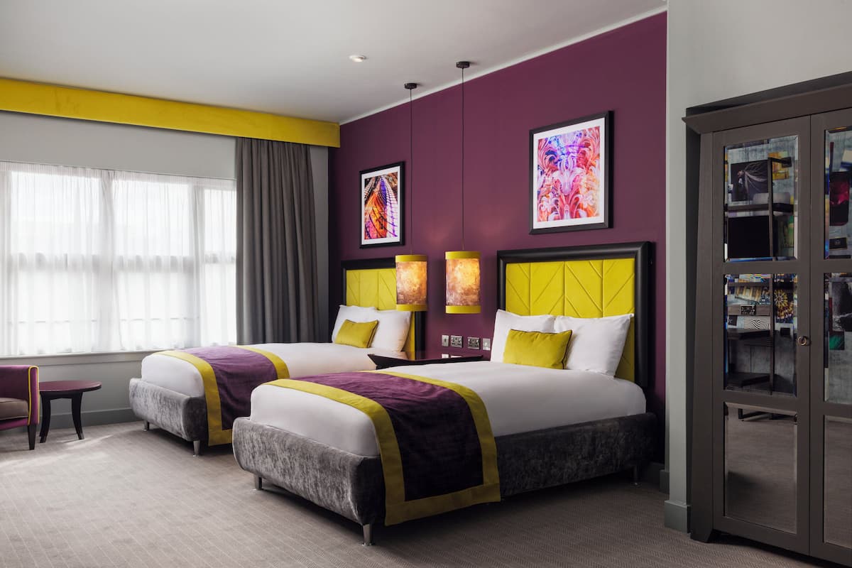 A hotel room with two beds, a large window with curtains, a desk, and a cabinet with glass doors.