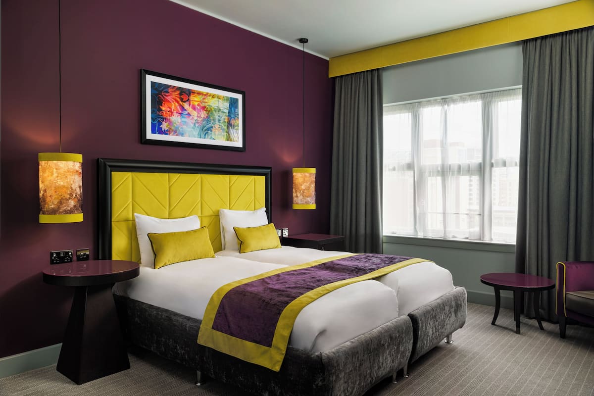 A modern hotel room with a large bed, two bedside tables, a small sofa, a window with curtains, and a city view.