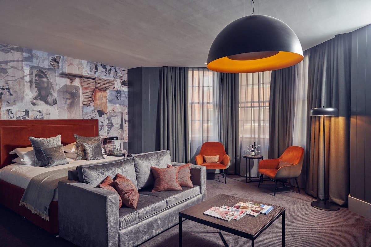 Moderni hotellihuone, jossa on suuri sänky, harmaa sohva, kaksi nojatuolia, sohvapöytä ja suuri riippuvalaisin.