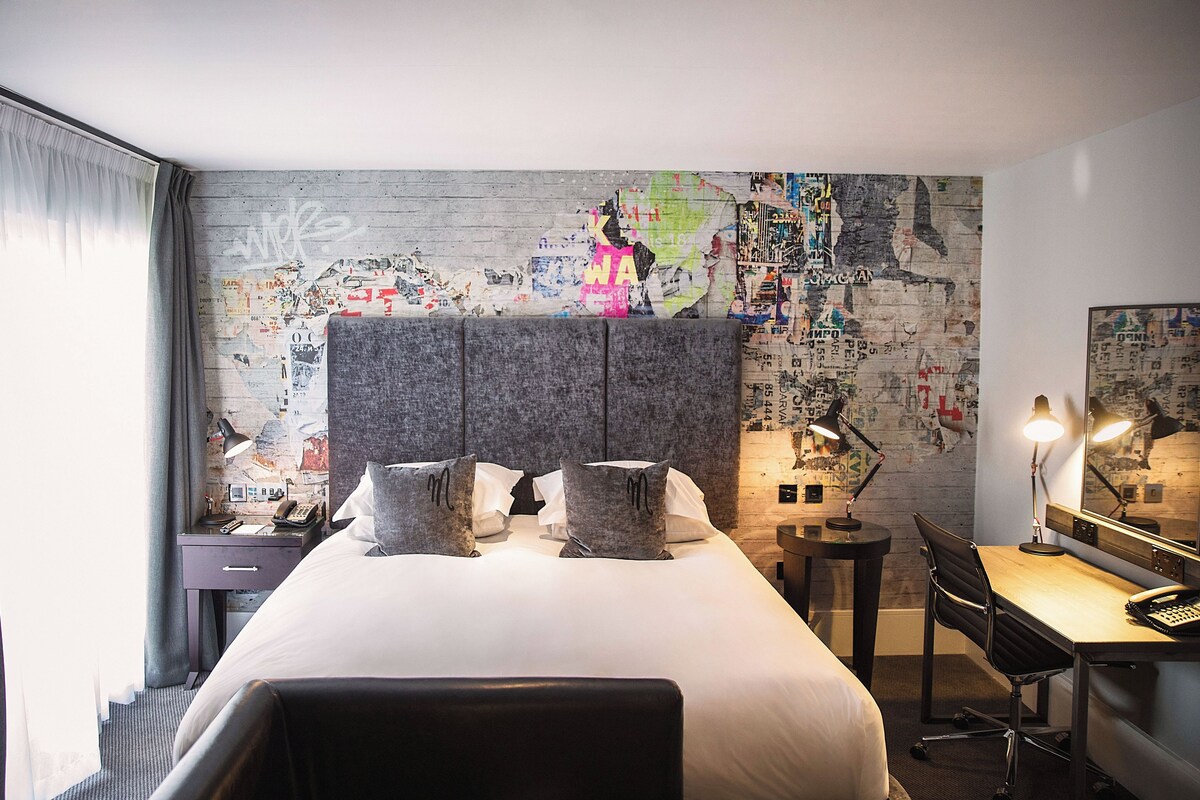 A hotel room with a large bed, a desk, a chair, a lamp, and a map-covered wall.