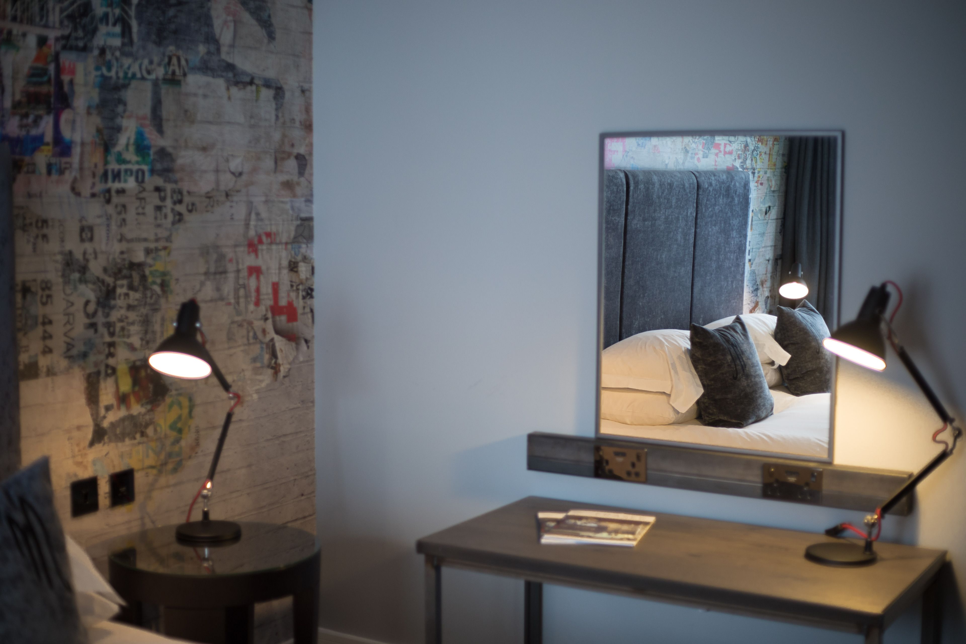 A hotel room with a bed, a desk, and a mirror. There are two lamps on the desk and a map on the wall.