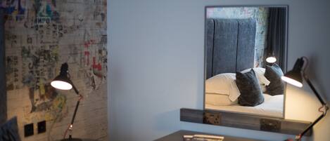 A hotel room with a bed, a desk, and a mirror. There are two lamps on the desk and a map on the wall.
