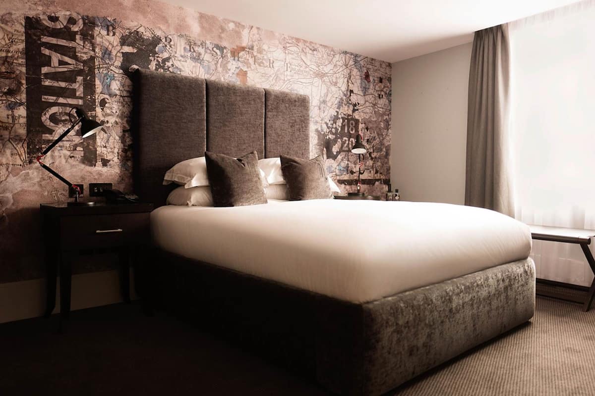 Moderni hotellihuone, jossa on suuri sänky, työpöytä ja karttoja esittävä sängynpääty.