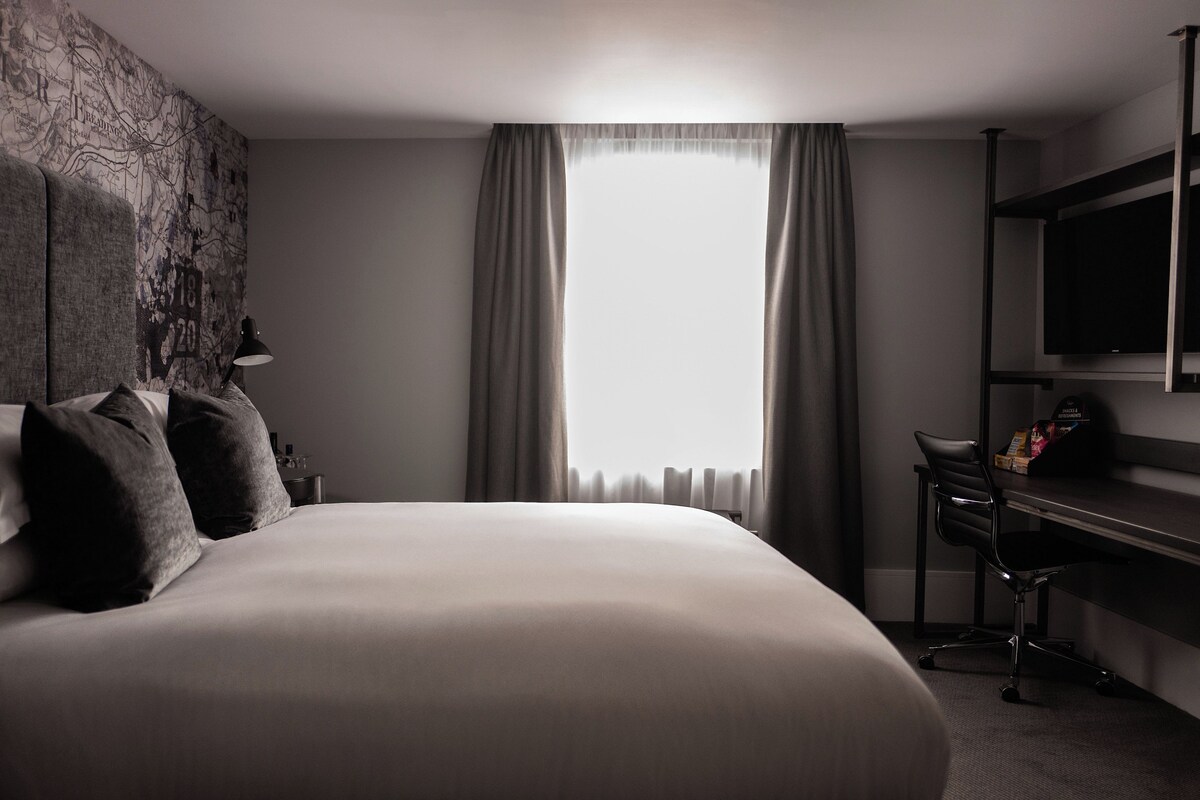 A hotel room with a large bed, a desk with a chair, a TV mounted on the wall, and a window with curtains.