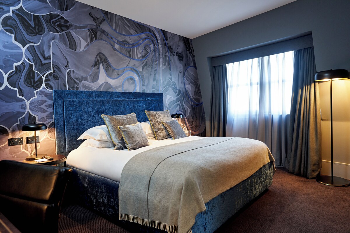 A modern hotel room with a large bed, a headboard, a desk, and a lamp.