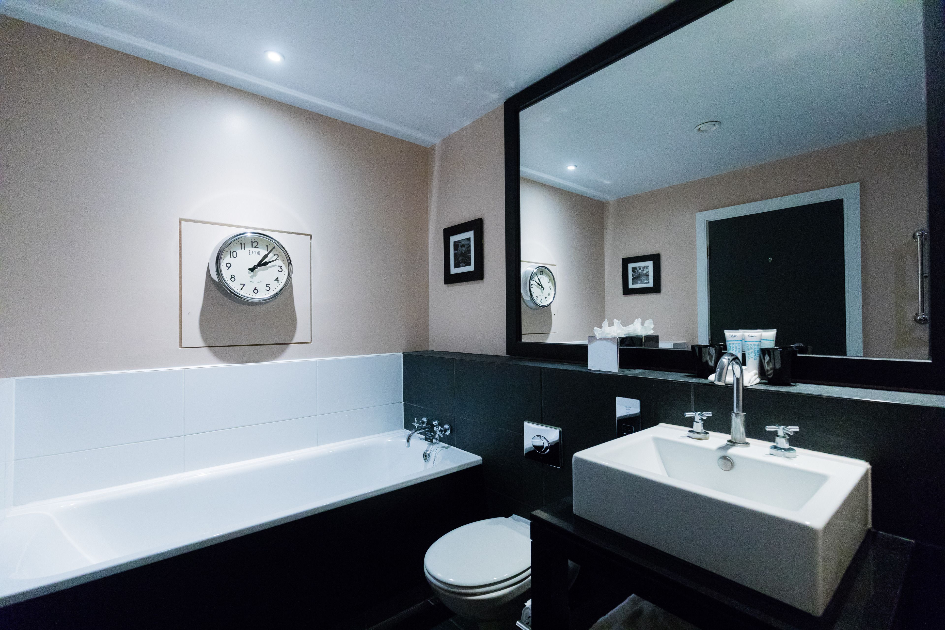 A modern bathroom with a bathtub, a sink, and a toilet. There are two clocks on the wall and a large mirror above the sink.