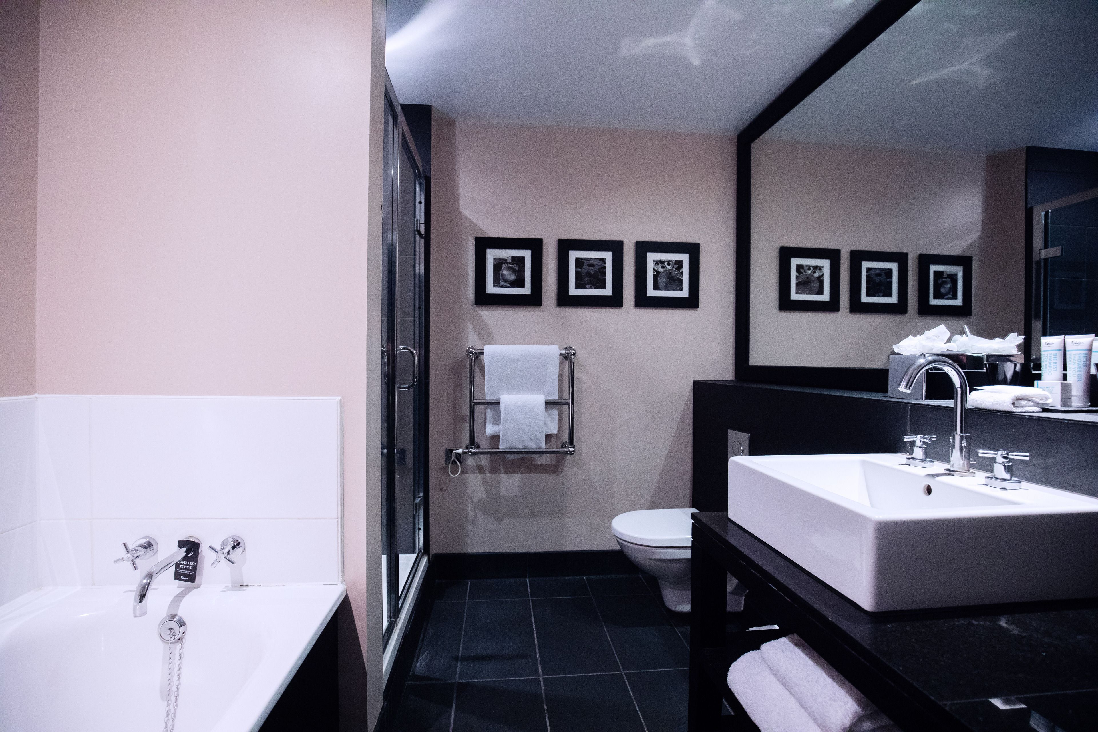 Een moderne badkamer met een bad, toilet en wastafel. Er hangen afbeeldingen in lijsten aan de muur en is een handdoekrek aanwezig.