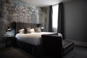 A modern hotel room with a large bed, a bedside table with a lamp, a dark-colored armchair, and a large window with curtains.