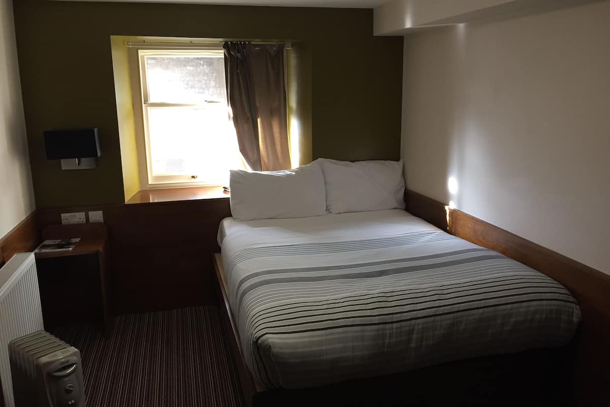 Une chambre d’hôtel avec un lit, une fenêtre avec des rideaux, une table de chevet et un téléviseur fixé au mur.