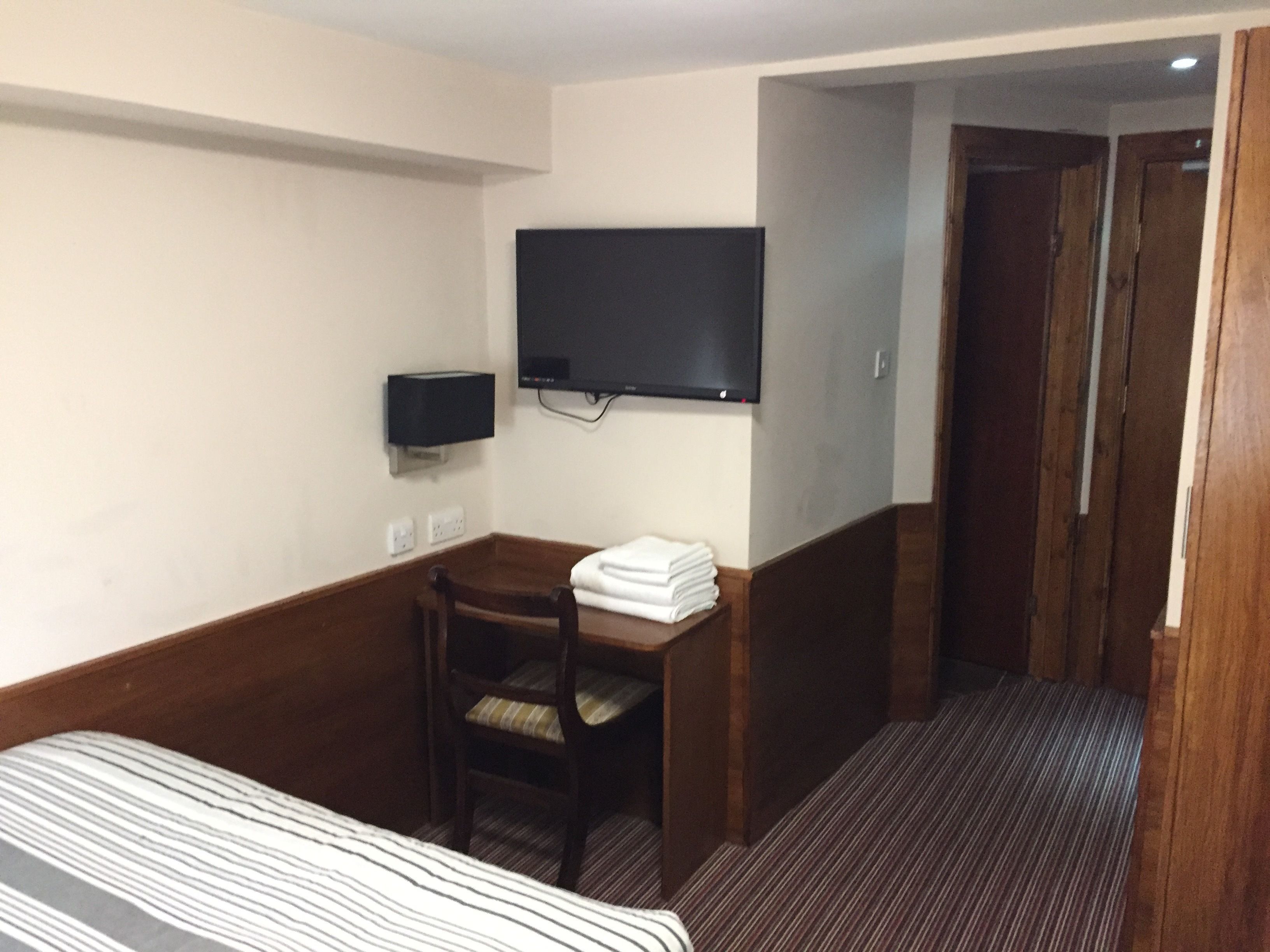 Habitación de hotel con cama, escritorio, silla, televisor montado en la pared y puerta que da a otra habitación.